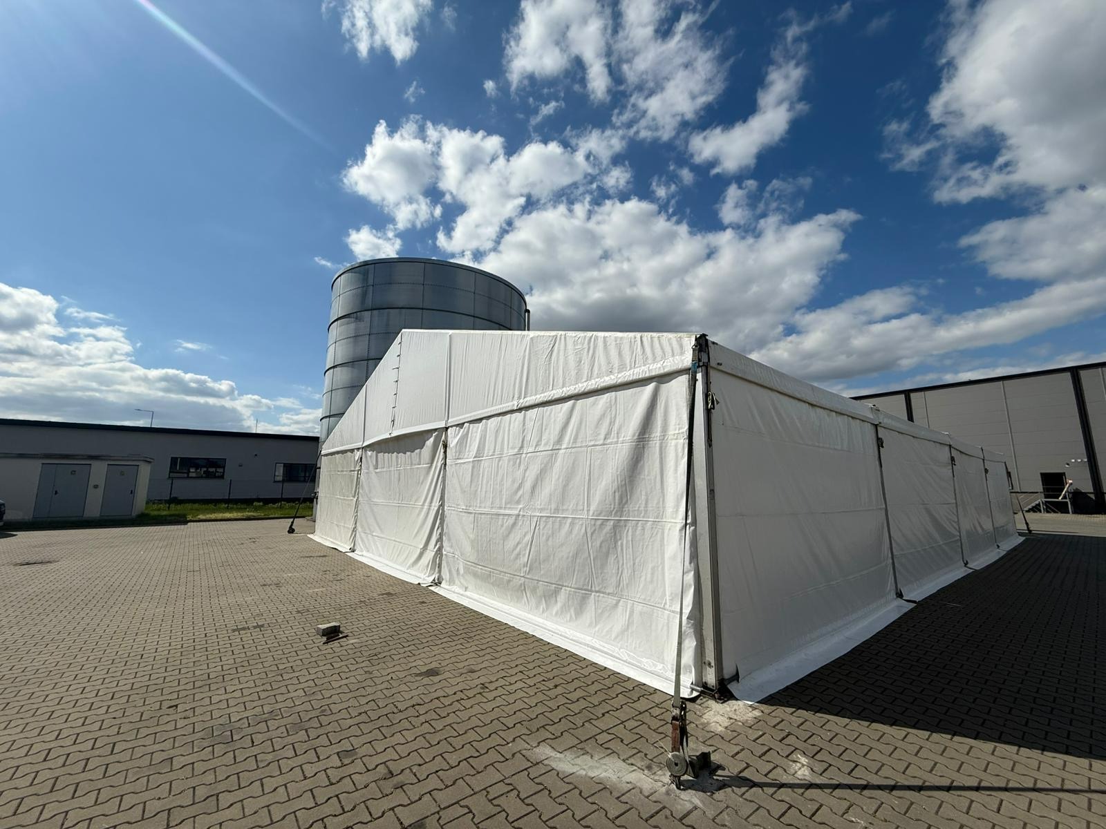 Rozłożony biały namiot plenerowy na kostce brukowej, z widocznym zbiornikiem przemysłowym w tle, pod błękitnym niebem z chmurami.