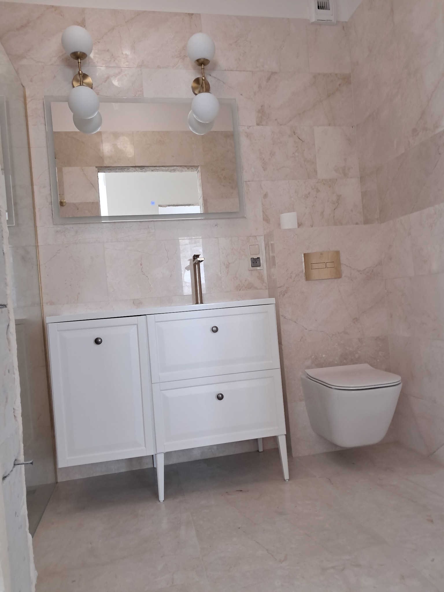 Elegancka łazienka w beżowych marmurach: biała szafka z umywalką, wisząca toaleta, lustro z oświetleniem. Minimalistyczny design, widoczne detale wykończenia.