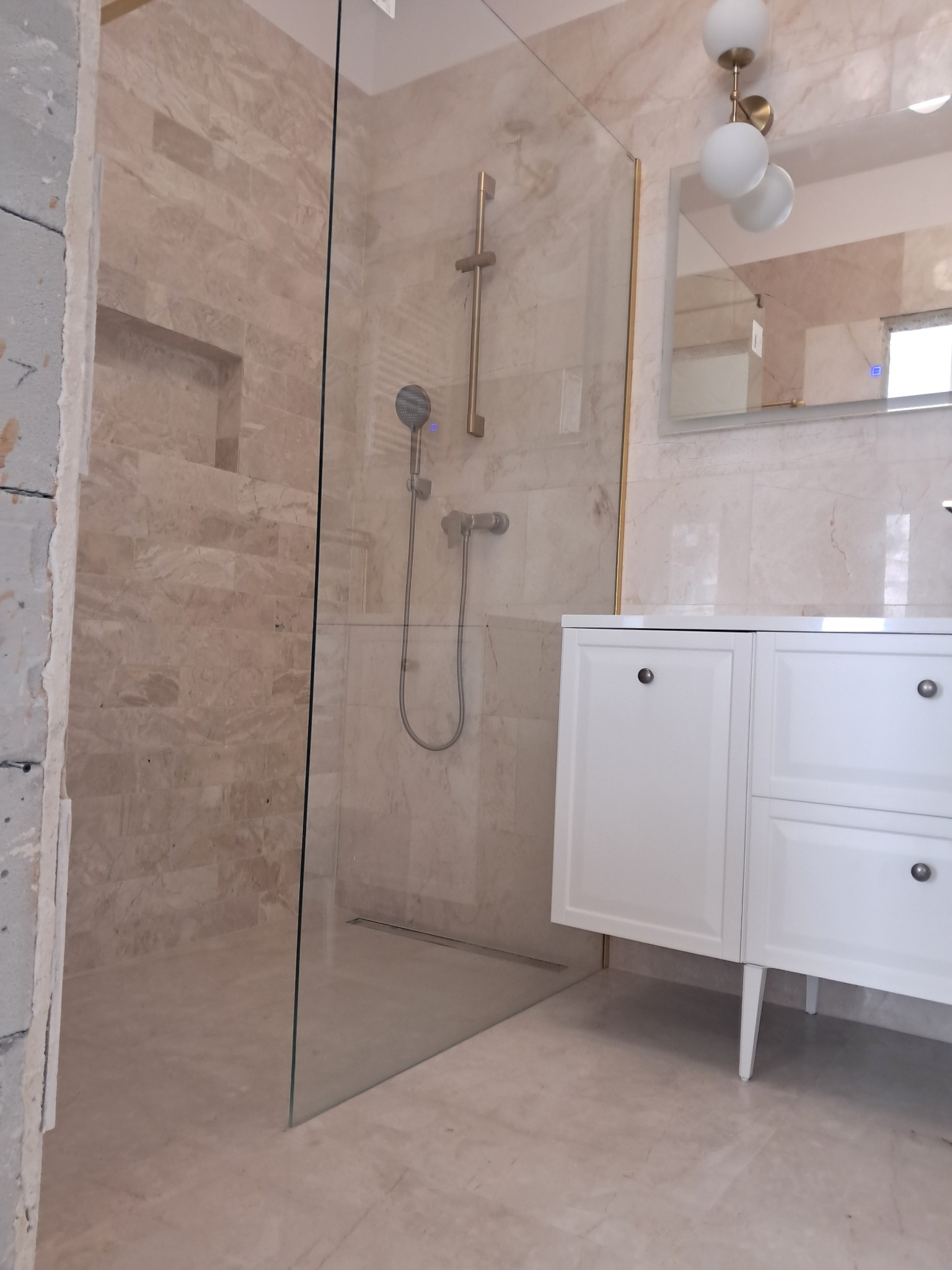 Wykończona kabina prysznicowa ze szklaną ścianką i armaturą w odcieniach szarości, umywalka z szafką w stylu klasycznym, beżowe płytki imitujące marmur.