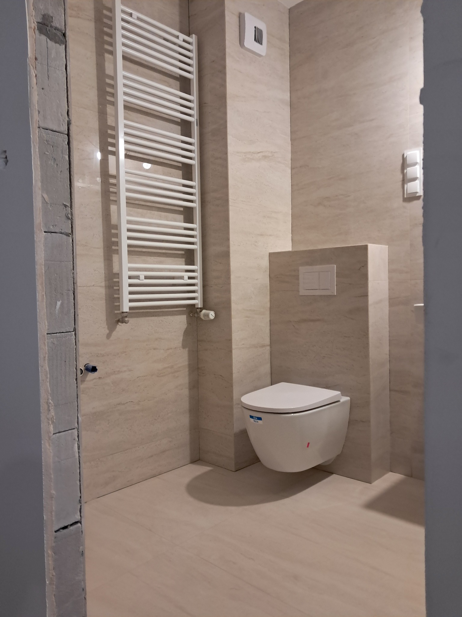 Nowoczesna łazienka po remoncie: beżowe płytki imitujące kamień, podwieszana toaleta i biały grzejnik drabinkowy. Widoczny fragment surowej ściany przy wejściu.