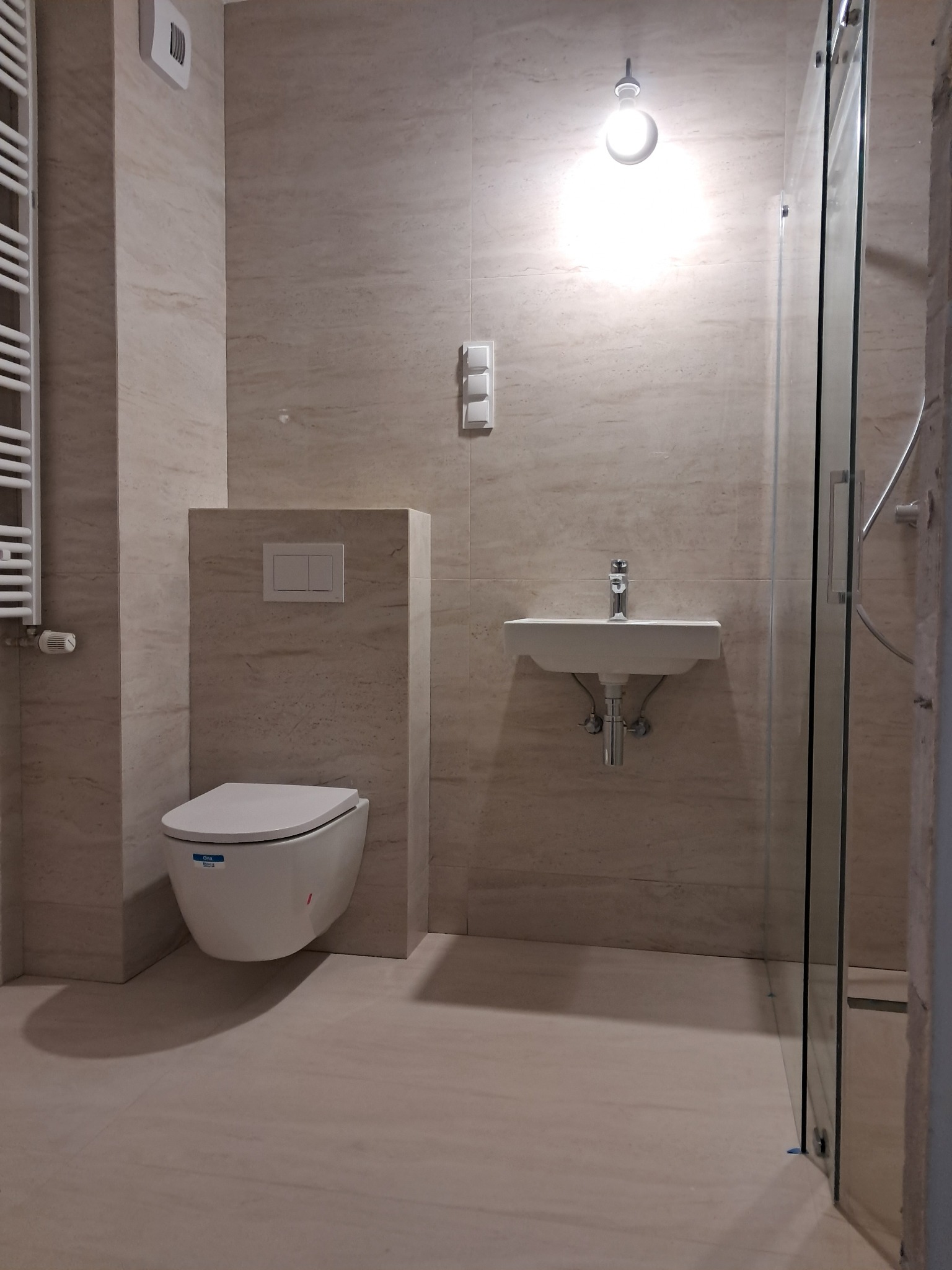 Nowoczesna łazienka z beżowymi płytkami imitującymi marmur, wisząca toaleta z zabudową, umywalka na ścianie i kabina prysznicowa z przezroczystego szkła.