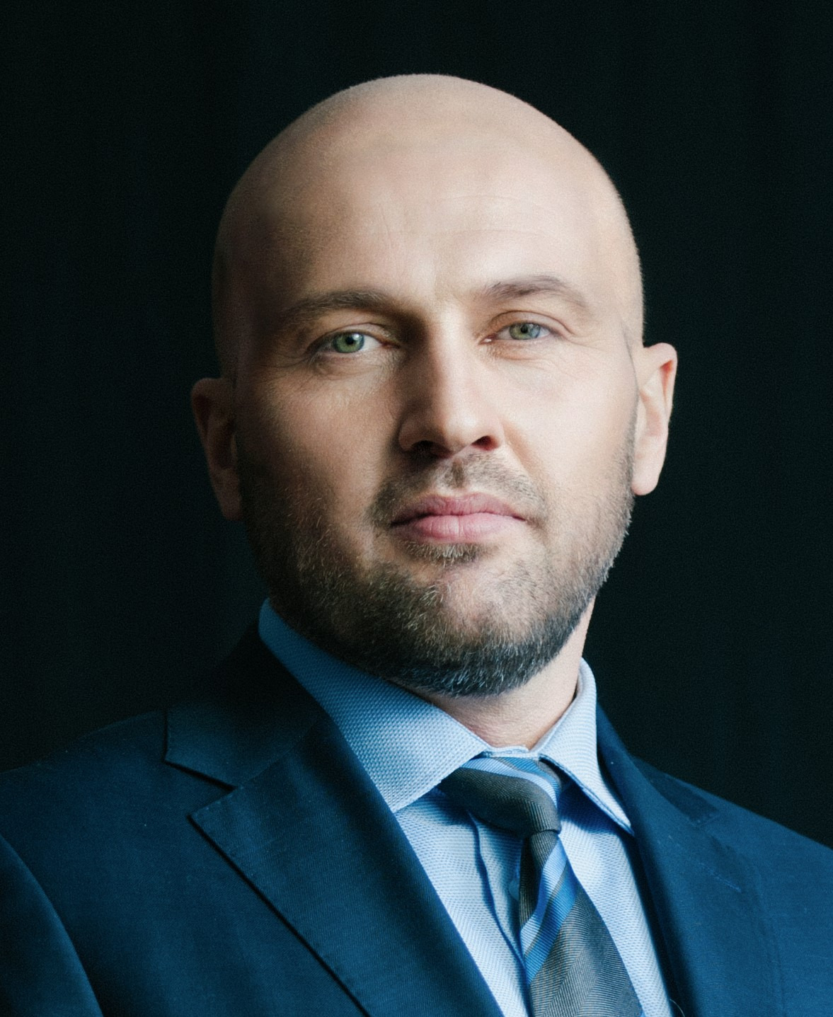 Portret mężczyzny w garniturze na ciemnym tle. Krótkie włosy, broda, niebieska koszula i krawat. Profesjonalny wygląd, bezpośrednie spojrzenie w obiektyw.