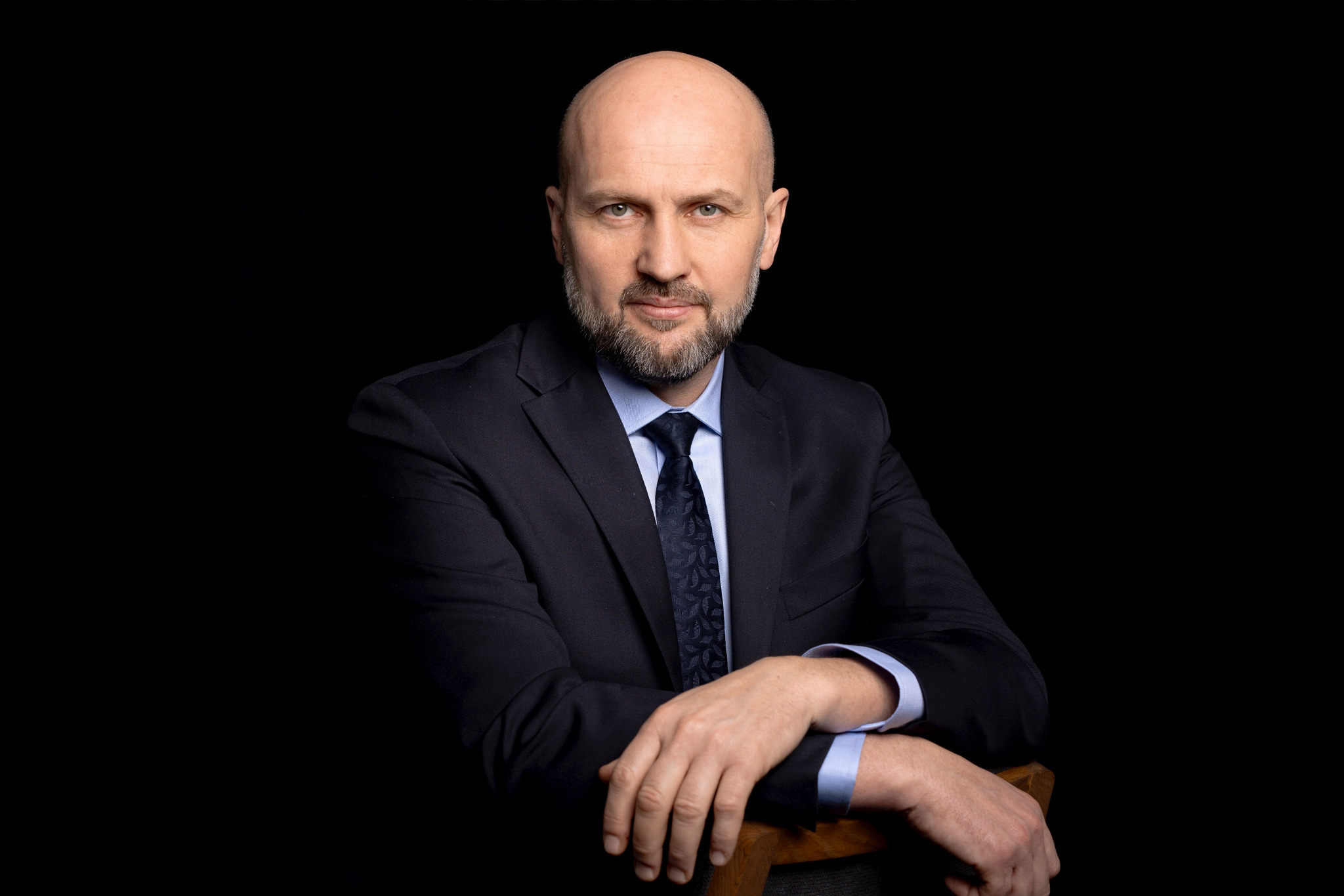 Portret biznesowy mężczyzny z brodą w ciemnym garniturze i błękitnej koszuli, opierającego ręce na oparciu krzesła na czarnym tle.