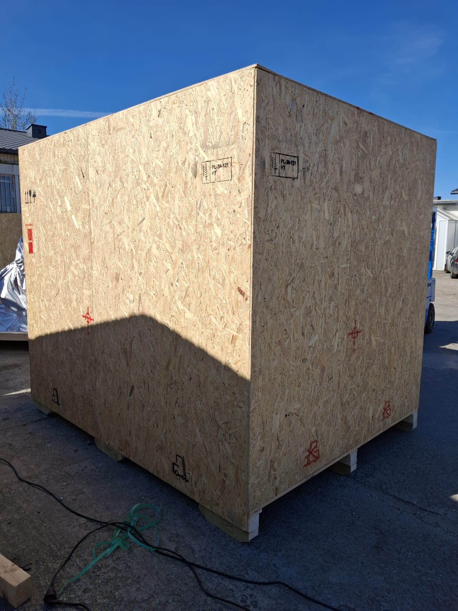 Duża, drewniana skrzynia transportowa na paletach, z widocznymi oznaczeniami, stojąca na zewnątrz w słoneczny dzień. W tle budynki i wózek widłowy.