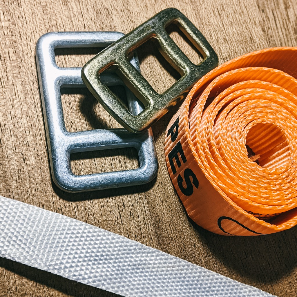 Metalowe klamry i zwinięty pomarańczowy pas tekstylny z napisem na tle drewnianej powierzchni. Elementy do pakowania i zabezpieczania ładunków.