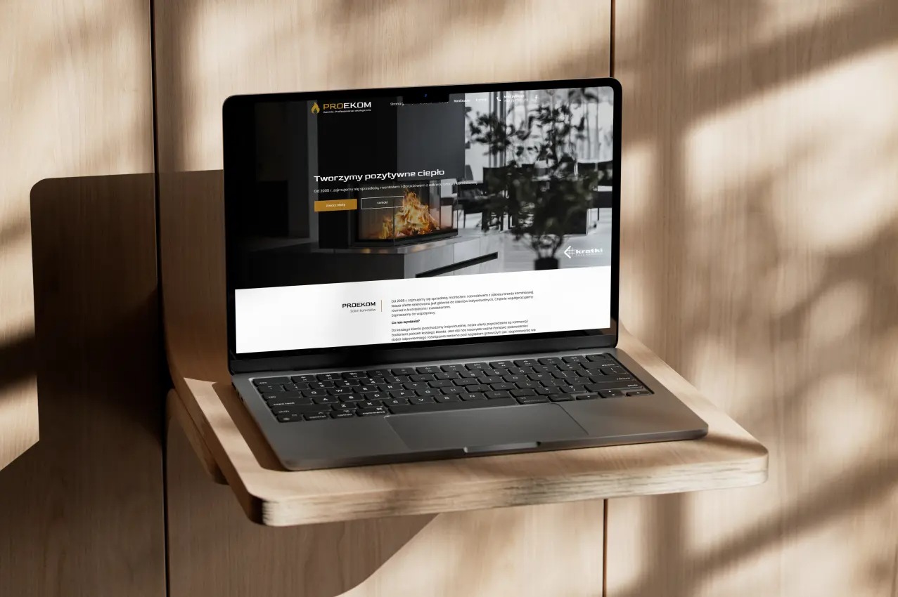Laptop prezentuje stronę www firmy Prodekom z kominkami, na tle drewnianej ściany z cieniem. Stonowana kolorystyka, nowoczesny design, ciepłe oświetlenie.
