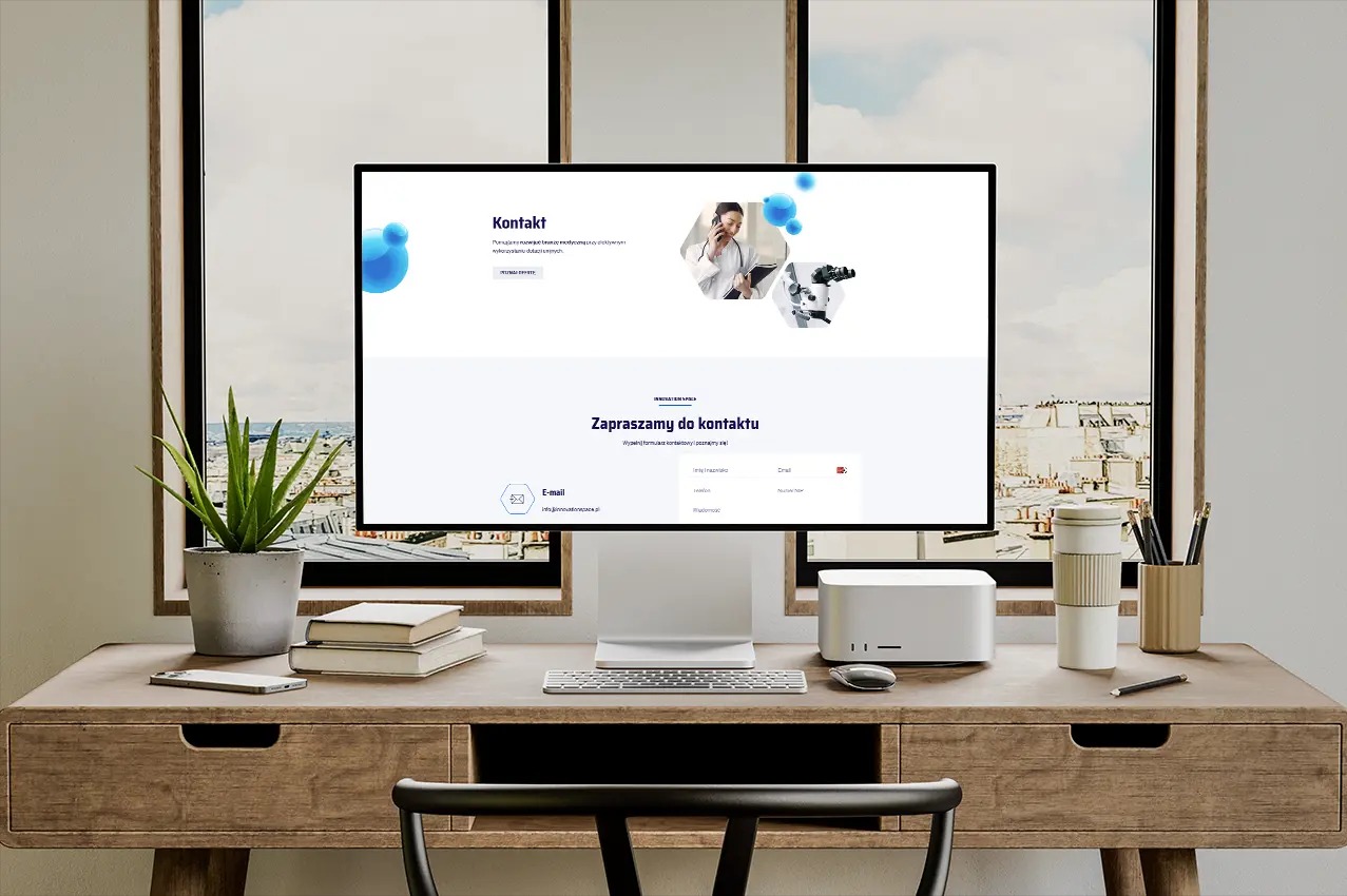 Monitor na biurku prezentuje stronę internetową z formularzem kontaktowym i motywem medycznym. W tle widoczne okna z widokiem na miasto.