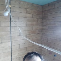 BOT - Wykończona łazienka z drewnopodobnymi płytkami na ścianach, niebieski sufit. Widoczna żarówka tymczasowa i fragment twarzy osoby robiącej zdjęcie.