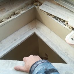 BOT - Ręka przytrzymuje fragment konstrukcji sufitu podwieszanego z płyt karton-gips, widoczne elementy mocujące i izolacja. Ujęcie z dołu.