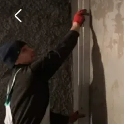 Pracownik w czerwonych rękawicach montuje metalowy profil pionowy na ścianie pokrytej szarą, chropowatą strukturą, przygotowując ją do dalszych prac wykończeniowych.