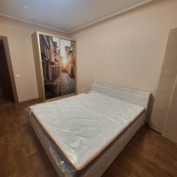 BOT - Jasne wnętrze w Opolu: łóżko z materacem w folii i szafa przesuwna z fototapetą uliczki. Świeżo wykończone mieszkanie, gotowe do umeblowania.