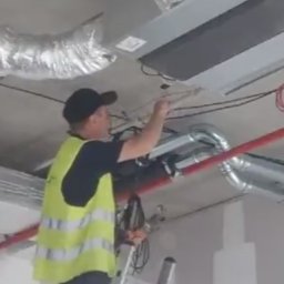 BOT - Pracownik w kamizelce na drabinie instaluje system wentylacyjny pod betonowym sufitem, widoczne rury i kable. Ujęcie od dołu.