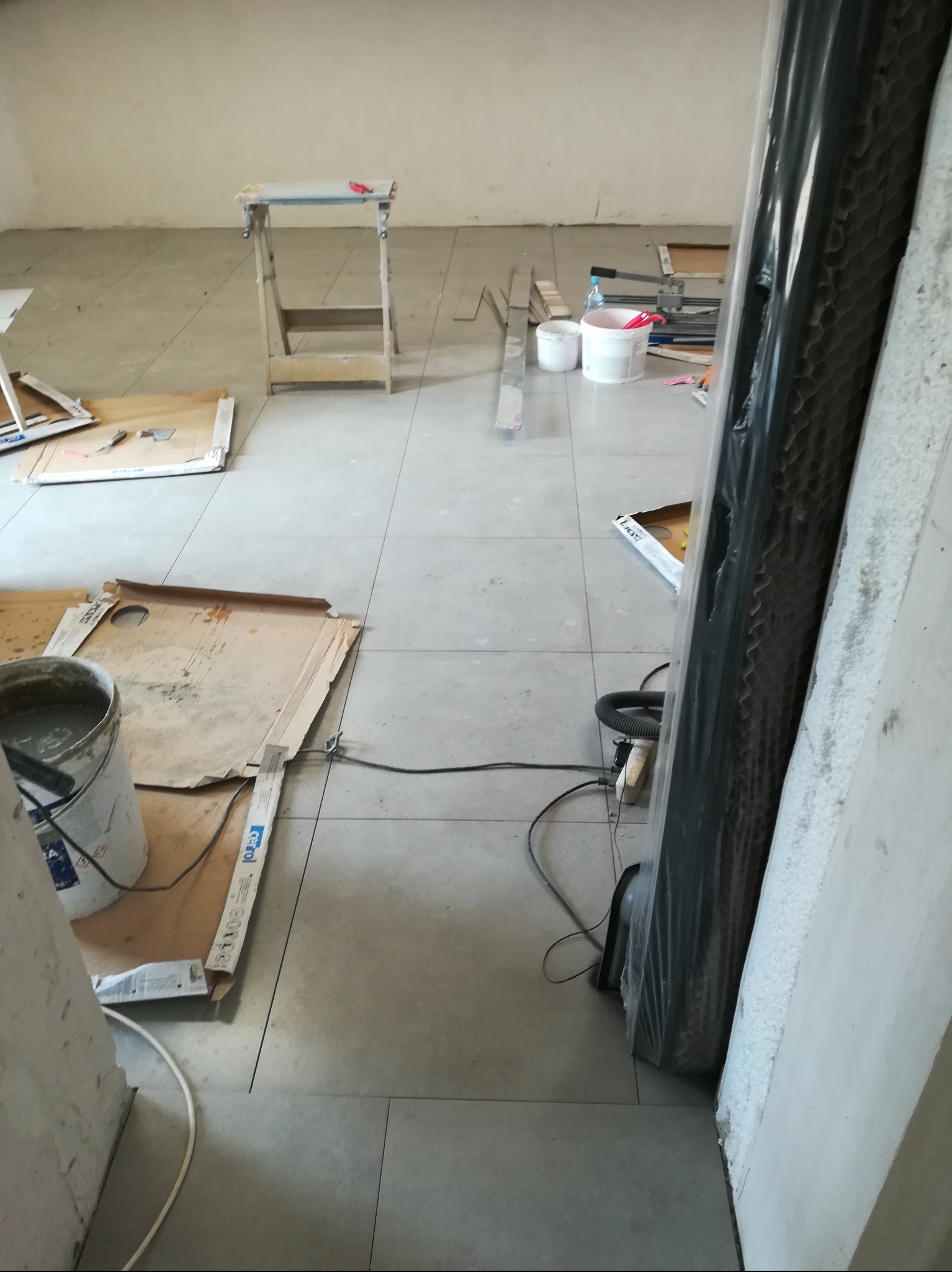 Jasnoszara podłoga z płytek w trakcie układania, widoczny stół roboczy, wiadro z zaprawą, paczki płytek i narzędzia glazurnicze. Całość w surowym wnętrzu.