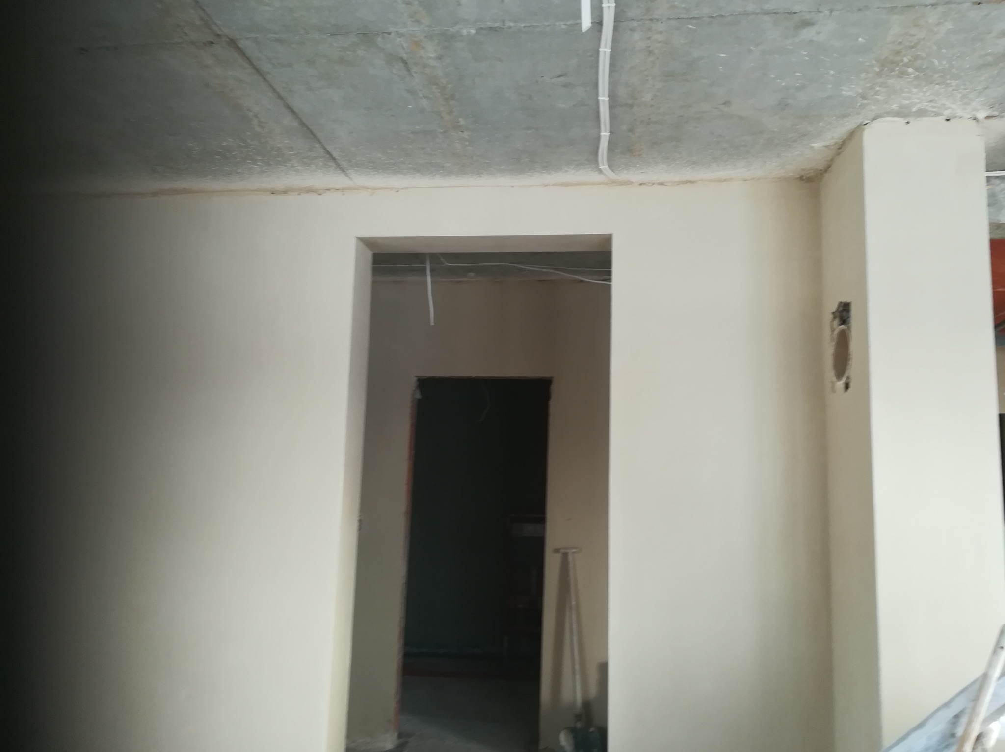 Wnętrze po gładziowaniu: gładkie ściany w jasnym odcieniu beżu, surowy betonowy sufit z widocznymi przewodami elektrycznymi, otwór drzwiowy w głębi.