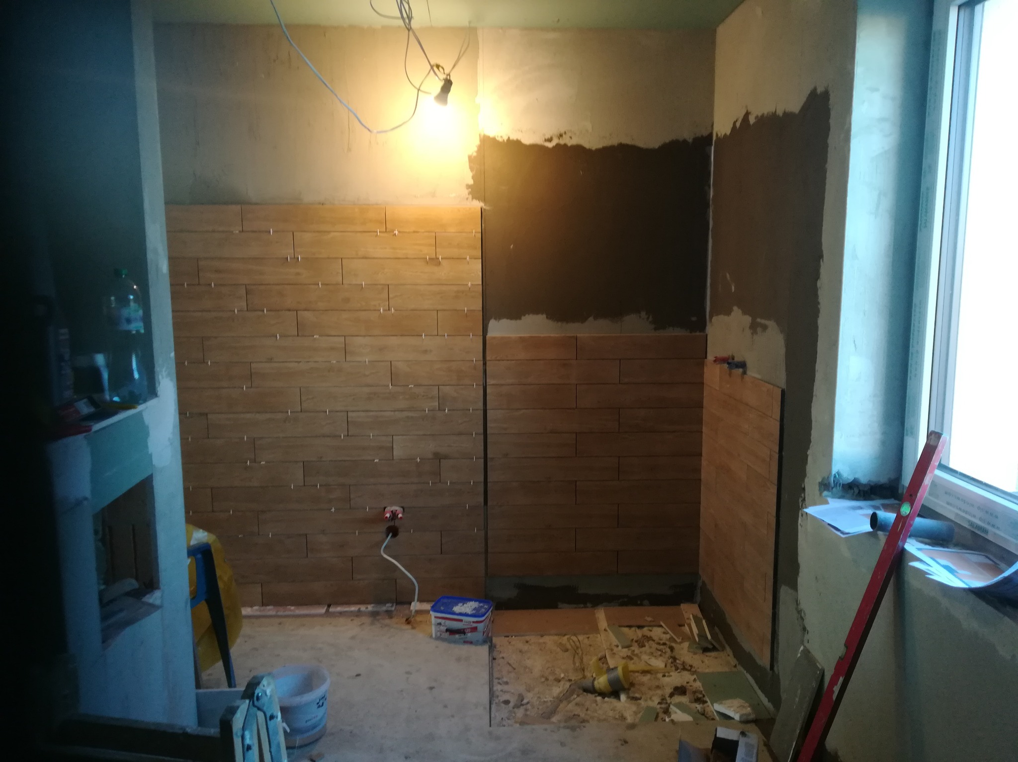 Częściowo wyłożona ściana płytkami imitującymi drewno w trakcie prac. Widoczne narzędzia i materiały budowlane. Oświetlenie halogenowe.