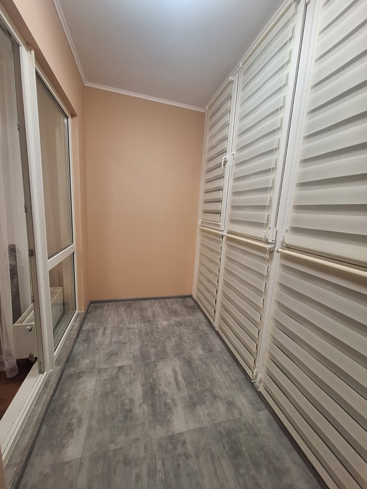 Wykończony balkon z szarymi płytkami podłogowymi, białymi oknami i roletami w kolorze ecru, tworzącymi nowoczesny i minimalistyczny wygląd.