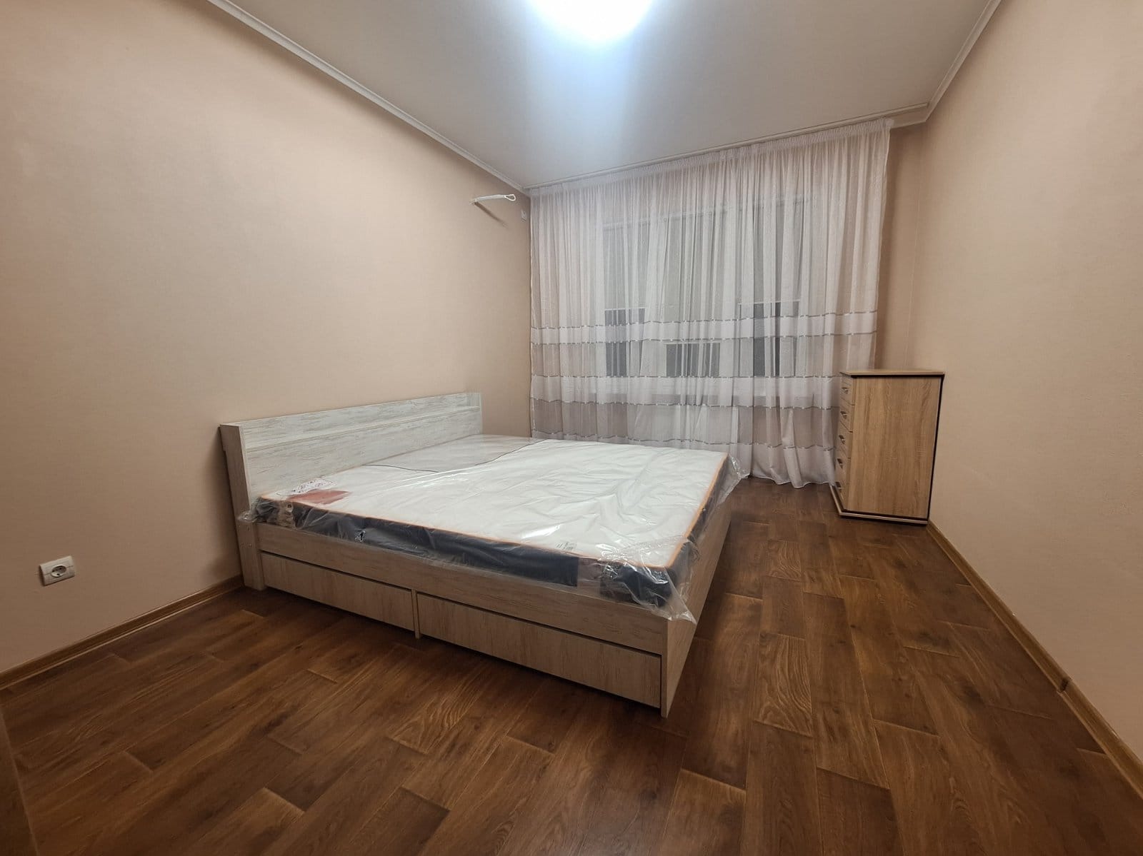 Jasna sypialnia po remoncie: drewniana podłoga, łóżko z materacem w folii, komoda, zasłony w oknie. Ściany w odcieniach beżu. Widok na nowo wykończone wnętrze.