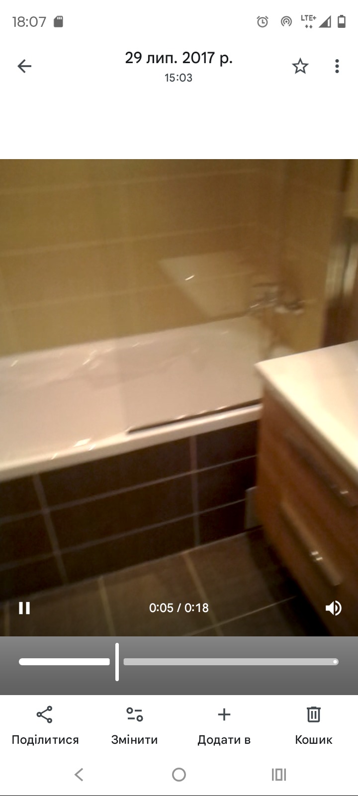 Wykończona łazienka z brązowymi płytkami, białą wanną z folią ochronną i drewnianą szafką pod umywalką. Widok z bliska, lekko rozmazany.