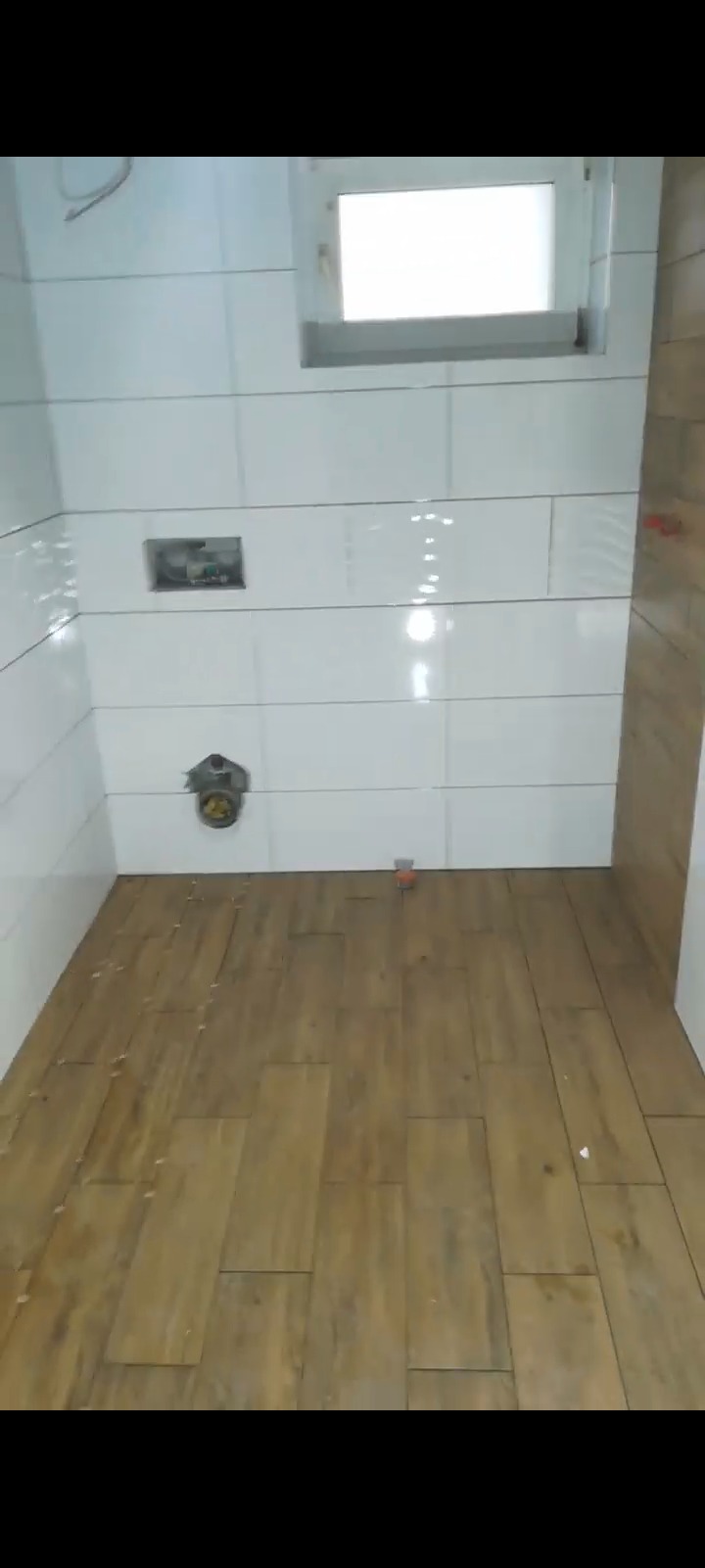 Białe, poziome płytki ścienne i drewnopodobna podłoga w trakcie wykańczania łazienki. Widoczne elementy instalacji i małe okno.