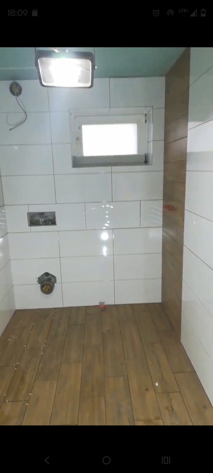 Wnętrze łazienki w trakcie prac: białe płytki ścienne, drewnopodobne płytki podłogowe, małe okno. Widoczna instalacja i oświetlenie robocze.