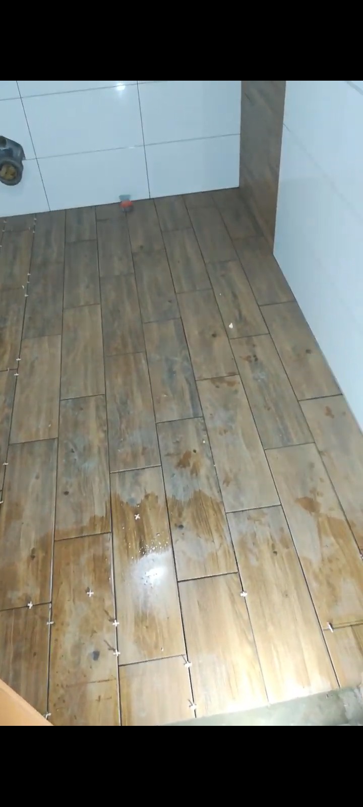 Układanie podłogowych płytek imitujących drewno w łazience, z użyciem krzyżyków dystansowych. Widoczne białe płytki na ścianie i fragment narzędzia glazurniczego.