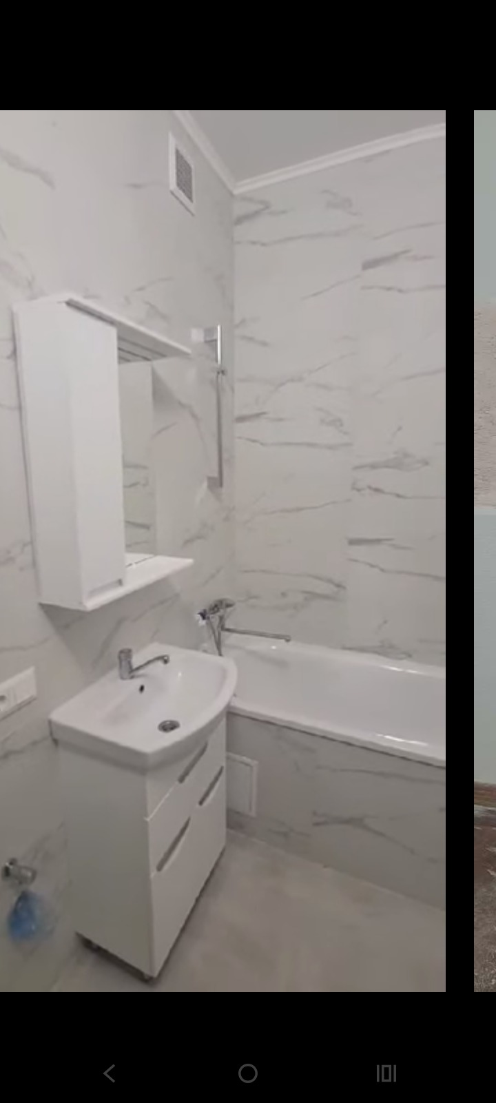 Biała łazienka z marmurowymi płytkami: umywalka z szafką, wanna i lustro z półką. Minimalistyczny design z delikatnym oświetleniem.