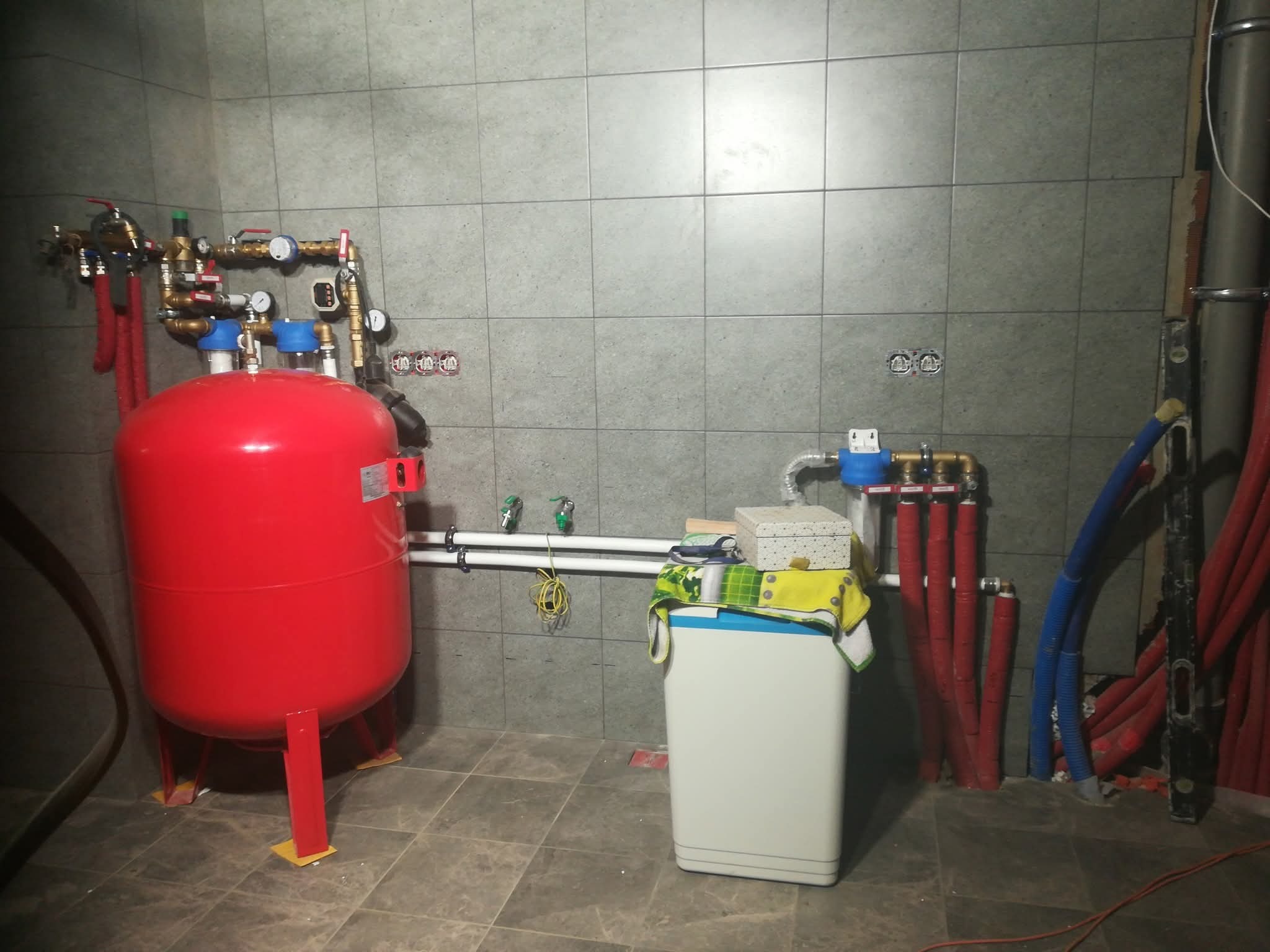 Zakończona instalacja centralnego ogrzewania z czerwonym zbiornikiem ciśnieniowym i szarymi płytkami na ścianie. Widoczne rury i zawory. Całość w pomieszczeniu technicznym.