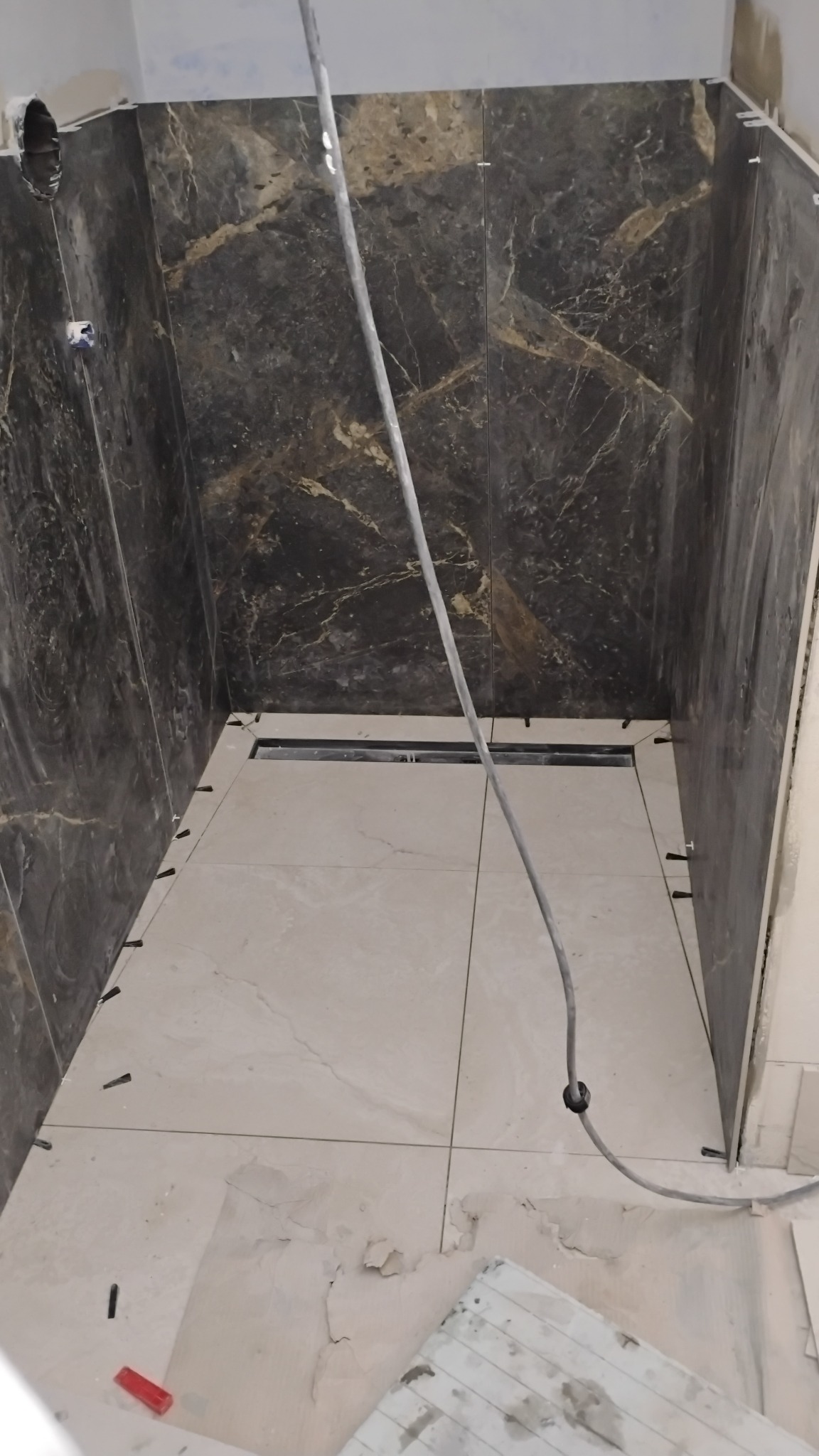 Wnętrze kabiny prysznicowej w trakcie wykańczania. Ułożone płytki podłogowe i ścienne z ciemnego marmuru. Widoczne krzyżyki dystansowe i odpływ liniowy.