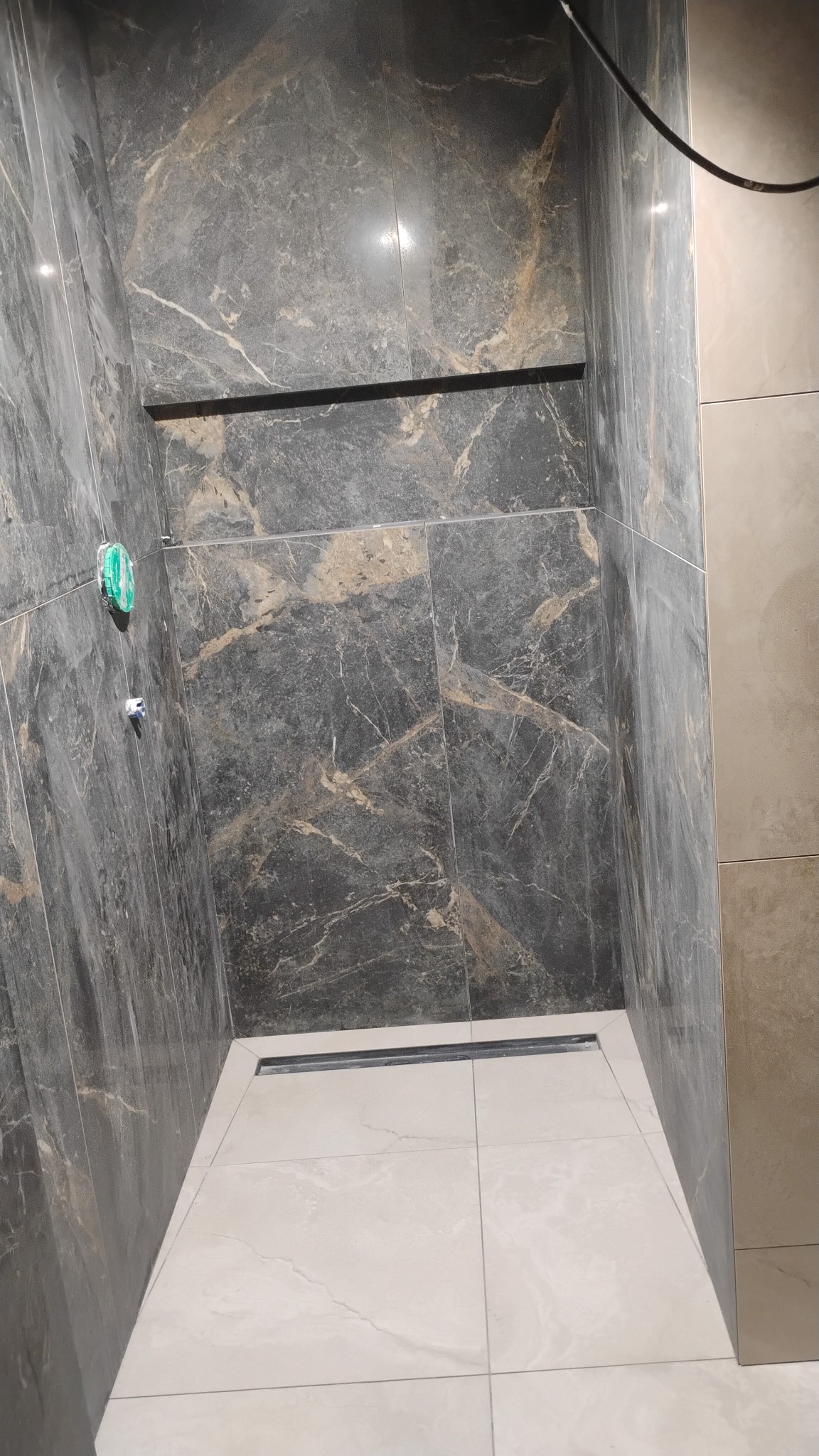 Wykończona kabina prysznicowa z ciemnymi płytkami imitującymi marmur na ścianach i jasnymi na podłodze. Widoczny odpływ liniowy i fragment instalacji.