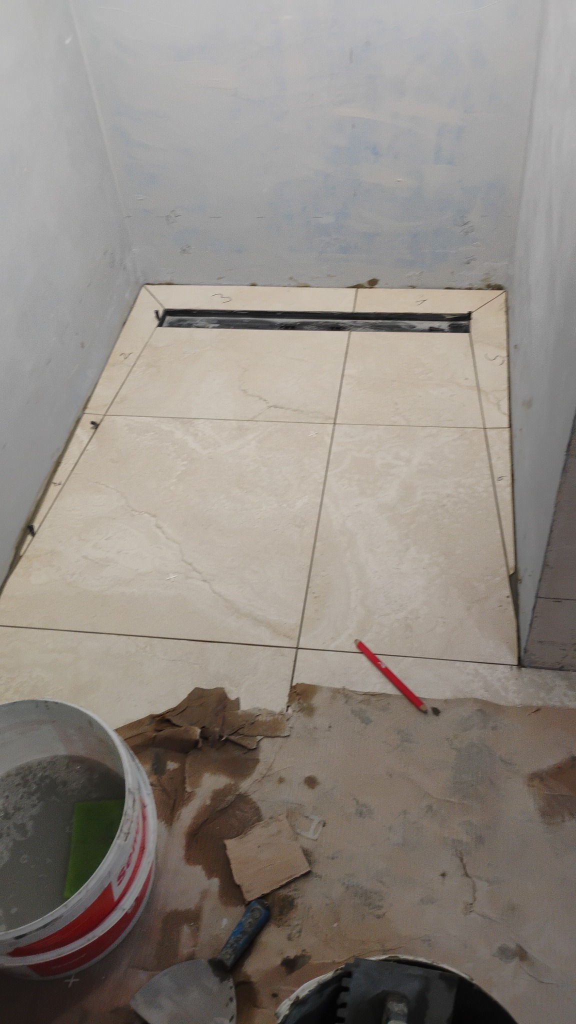 Układanie beżowych płytek podłogowych w łazience z odpływem liniowym. Widoczny ołówek, wiadro i narzędzia glazurnicze. Prace w toku, podłoże przygotowane pod dalszy etap.