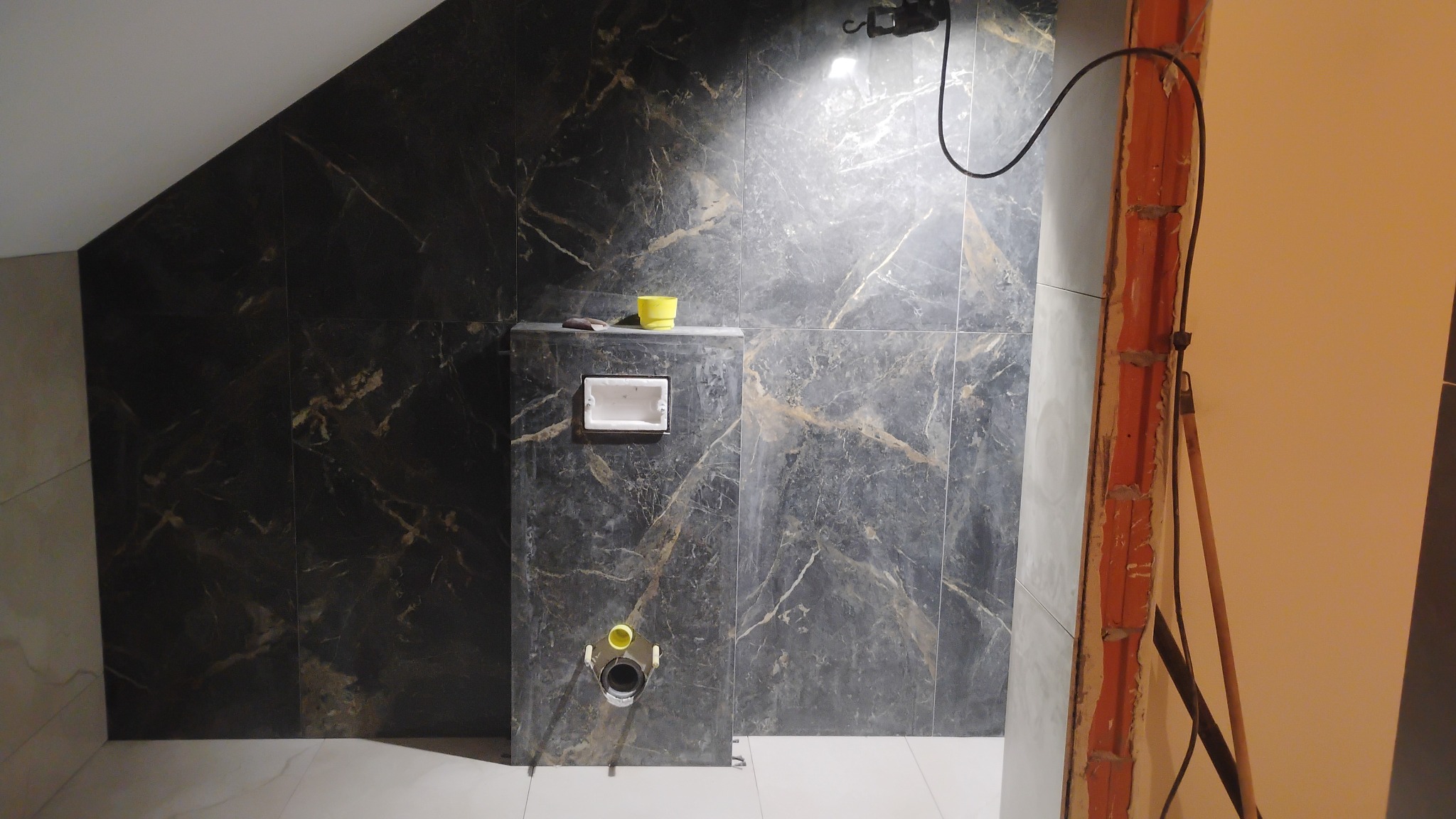 Częściowo wykończona łazienka z czarnymi płytkami imitującymi marmur na ścianach, widoczna instalacja podtynkowa WC i fragment ściany w trakcie prac wykończeniowych.