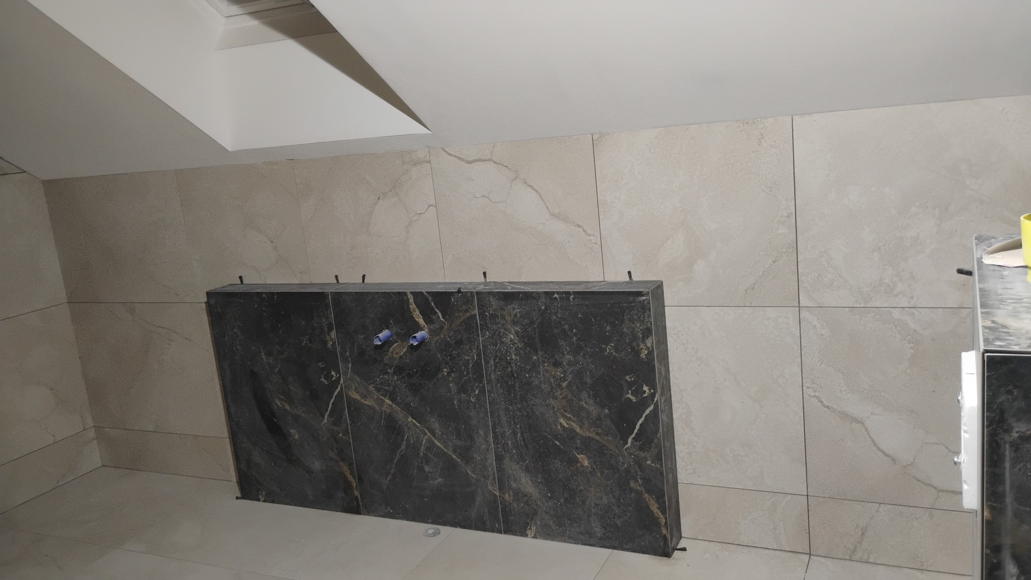 Montaż czarnych, marmurkowych płyt na ścianie w łazience. Widoczne elementy mocujące. Beżowe płytki na ścianach i podłodze. Ukośny sufit.