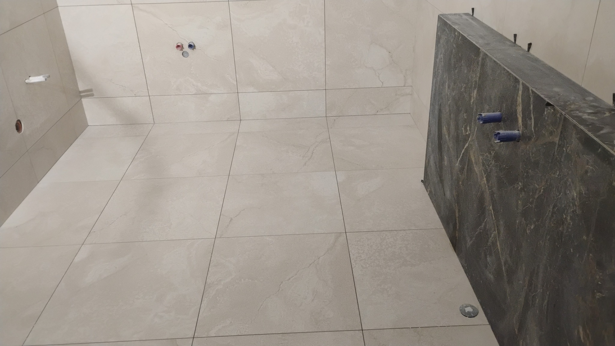 Wykończona łazienka z beżowymi płytkami podłogowymi i ściennymi. Widoczny fragment ściany z ciemnego kamienia z wystającymi rurami. Ujęcie z dołu.