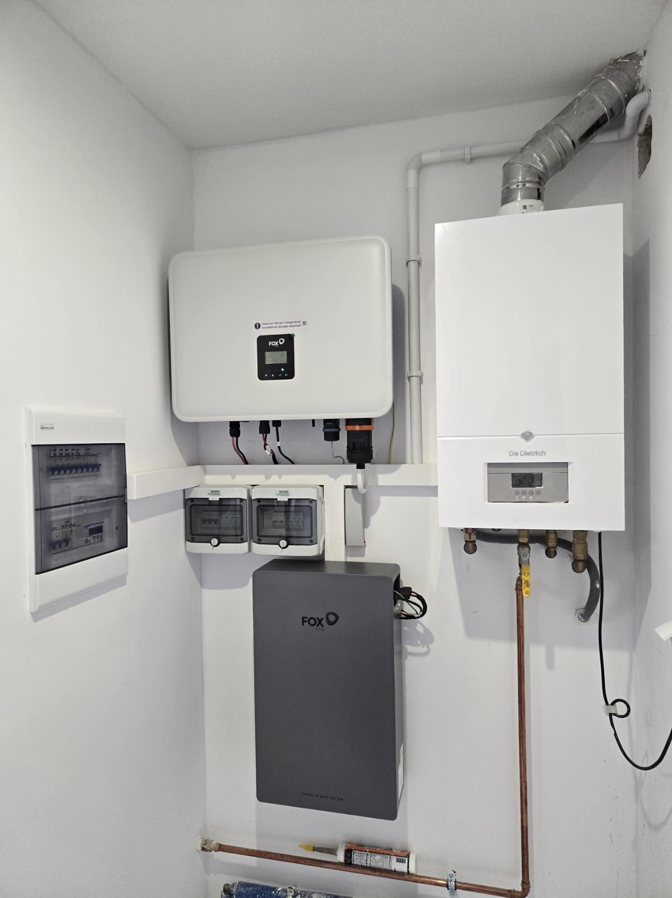 Kompletna instalacja: inwerter fotowoltaiczny Fox ESS, magazyn energii, licznik dwukierunkowy, rozdzielnia elektryczna i piec gazowy De Dietrich na białej ścianie.