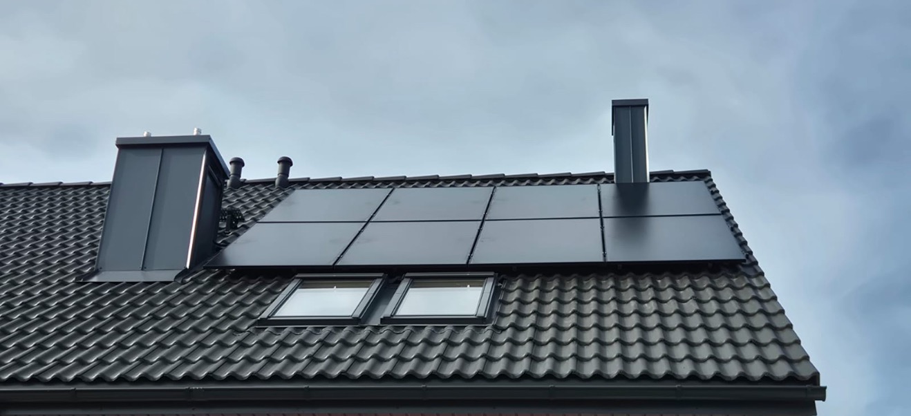 Czarny dach z panelami fotowoltaicznymi i oknami dachowymi, kominy w kolorze dachu, widok z dołu na tle pochmurnego nieba.