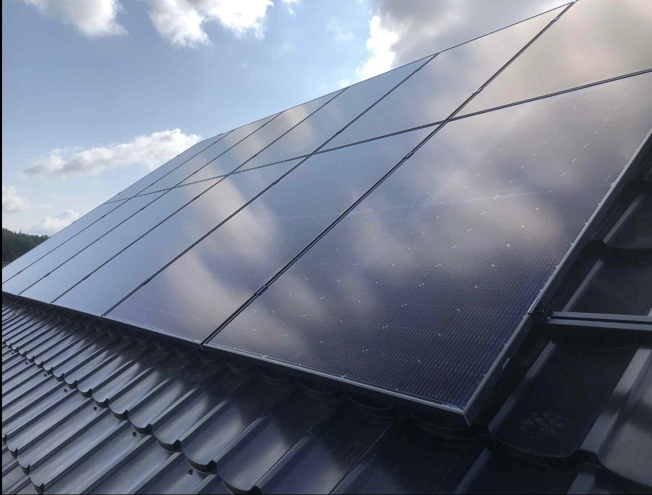 Panele fotowoltaiczne na ciemnym dachu z dachówki ceramicznej, odbijające błękit nieba i chmury. Widok pod kątem, na tle fragmentu lasu.