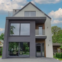 RAVEN S.C. KRZYSZTOF BOŚKA, SŁAWOMIR BŁASZCZAK - Nowoczesny dom z dużymi oknami i balkonem z szklaną balustradą. Elewacja w odcieniach szarości i bieli. Widoczne drzewa i błękitne niebo z chmurami.