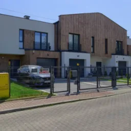 Nowoczesne segmenty mieszkalne z drewnianą elewacją, balkonami i garażami, oddzielone metalowym ogrodzeniem z bramą wjazdową. Na podjeździe stoi szary samochód.