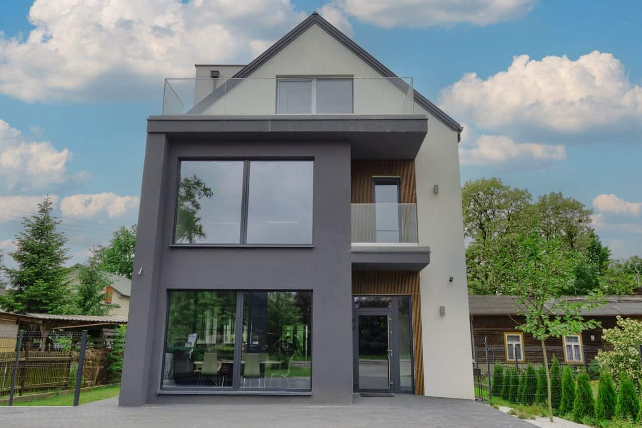 Nowoczesny dom z dużymi oknami i balkonem z szklaną balustradą. Elewacja w odcieniach szarości i bieli. Widoczne drzewa i błękitne niebo z chmurami.