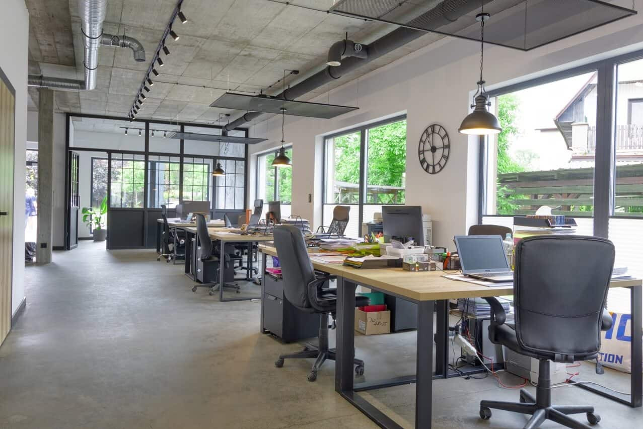 Nowoczesne biuro z widokiem na zewnątrz przez okna z czarnymi ramami. Widoczne biurka, krzesła obrotowe i betonowa podłoga.