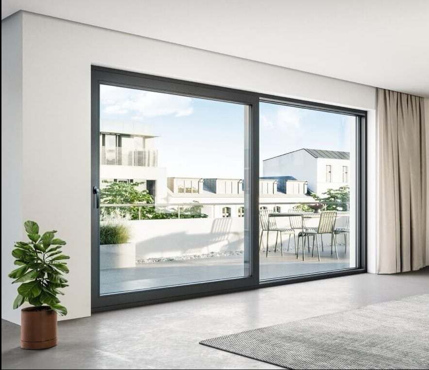 Nowoczesne drzwi przesuwne w minimalistycznym wnętrzu z widokiem na taras i budynki w tle. Szara rama, roślina doniczkowa i dywan dodają przytulności.
