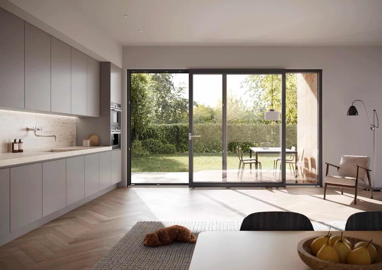 Nowoczesne, duże drzwi przesuwne z ciemną ramą, otwarte na ogród, w jasnym wnętrzu z drewnianą podłogą i szarymi meblami kuchennymi. W tle widoczny stół i krzesła.