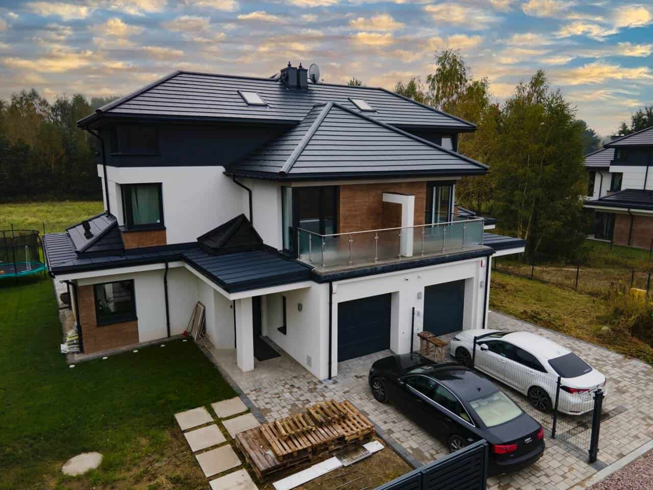 Nowoczesny dom z balkonem z balustradą ze szkła, dwustanowiskowym garażem i podjazdem z kostki. Widoczne okna i dach z dachówkami.