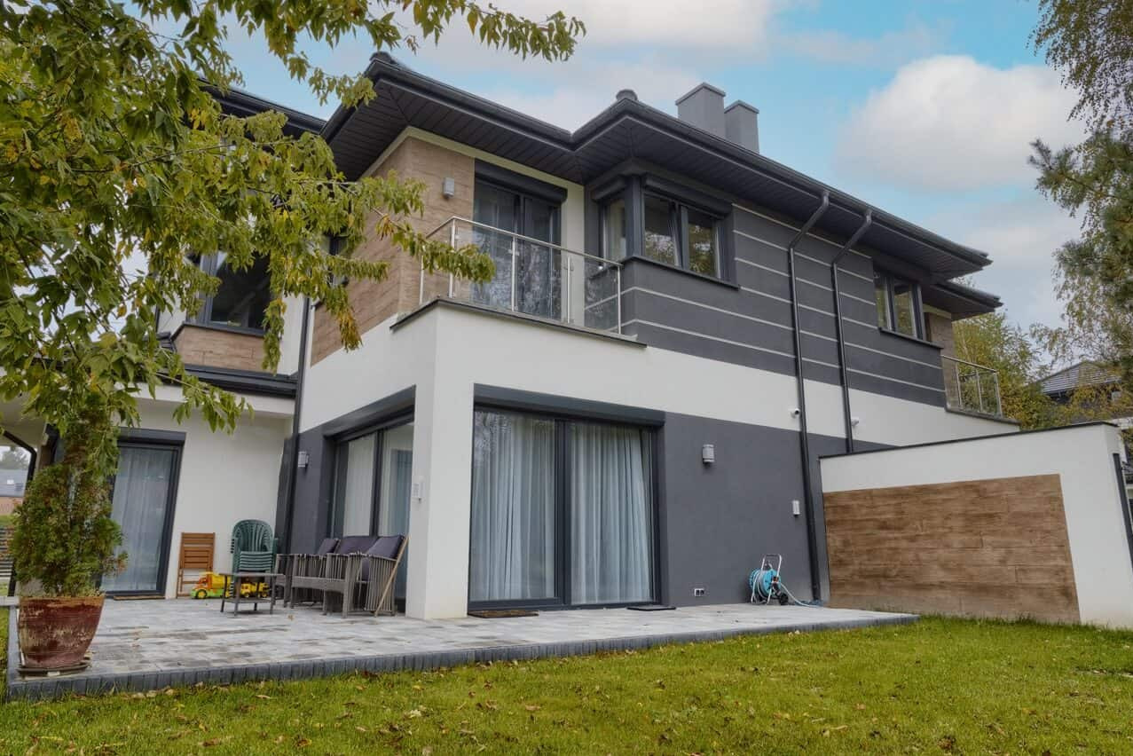 Nowoczesny dom jednorodzinny z szarymi oknami PCV i roletami zewnętrznymi, widok z ogrodu na taras z przesuwnymi drzwiami balkonowymi. Detal: zieleń drzew w kadrze.