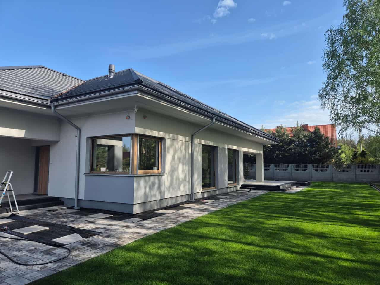 Nowoczesny dom jednorodzinny z dużymi oknami PCV, widok z zewnątrz na trawnik i taras. Widoczne elementy wykończeniowe i detale architektoniczne.