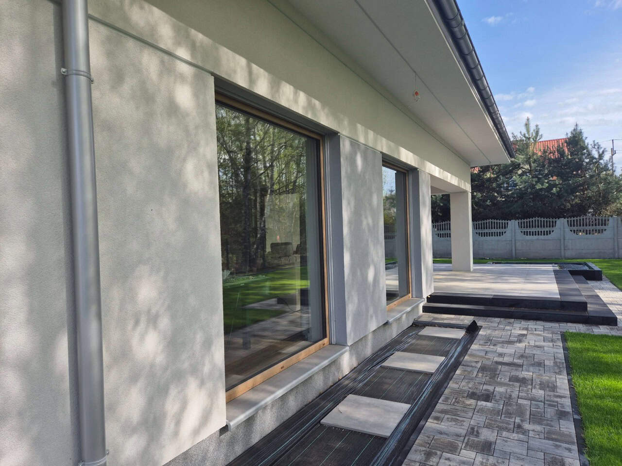 Nowoczesny dom z dużymi oknami PCV z drewnianą ramą, widok z zewnątrz. Elegancki design, jasna elewacja i zadbany ogród. Widoczne detale wykończenia.