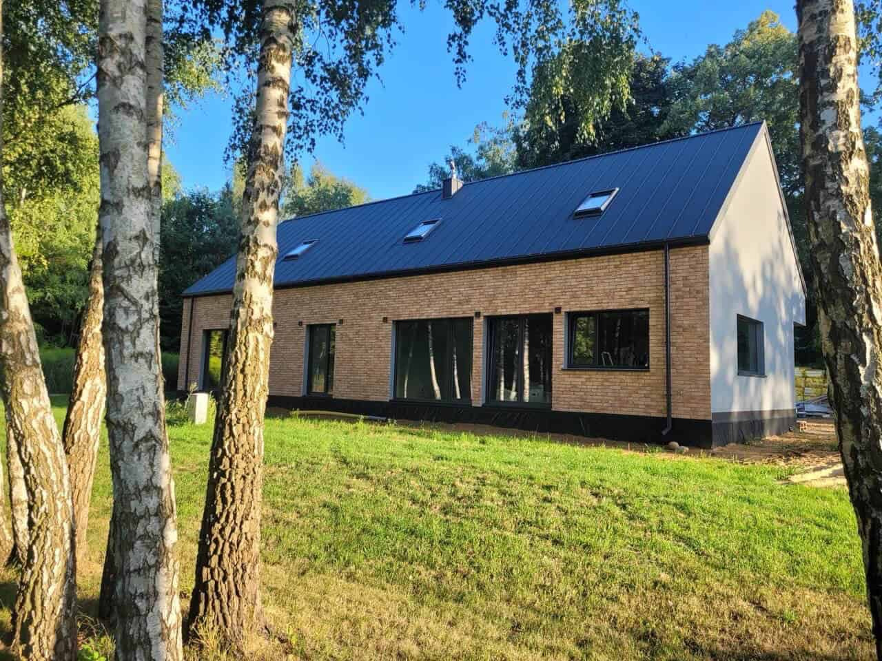 Nowoczesny dom z ceglaną elewacją i ciemną stolarką okienną, widoczny zza drzew. Dach pokryty blachą, z oknami dachowymi. Jasne, słoneczne otoczenie.