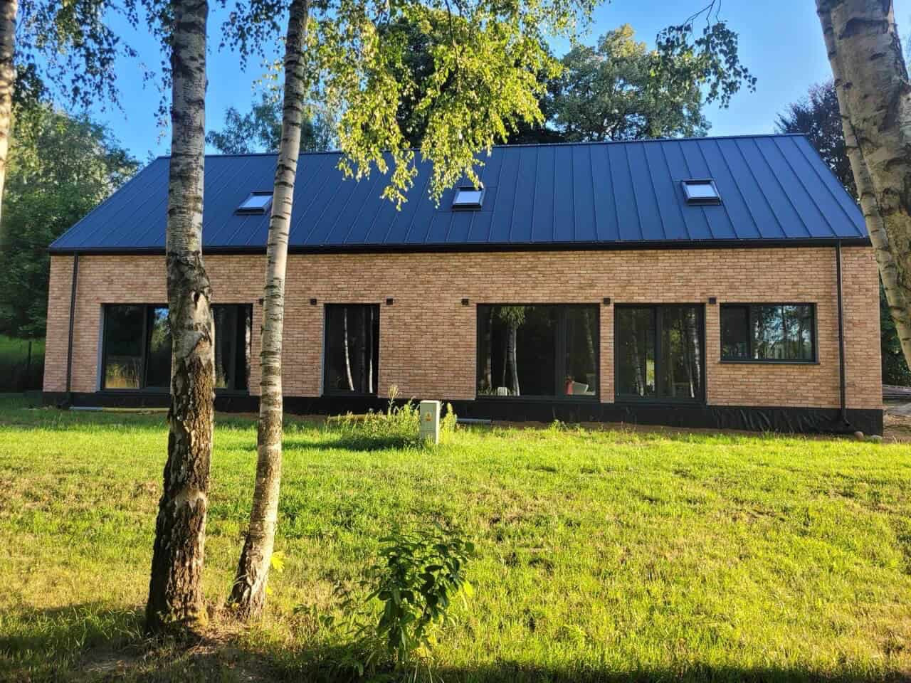 Nowoczesny dom z cegły klinkierowej z ciemną blachodachówką i czarnymi oknami, widoczny z perspektywy trawnika, z brzozami na pierwszym planie.