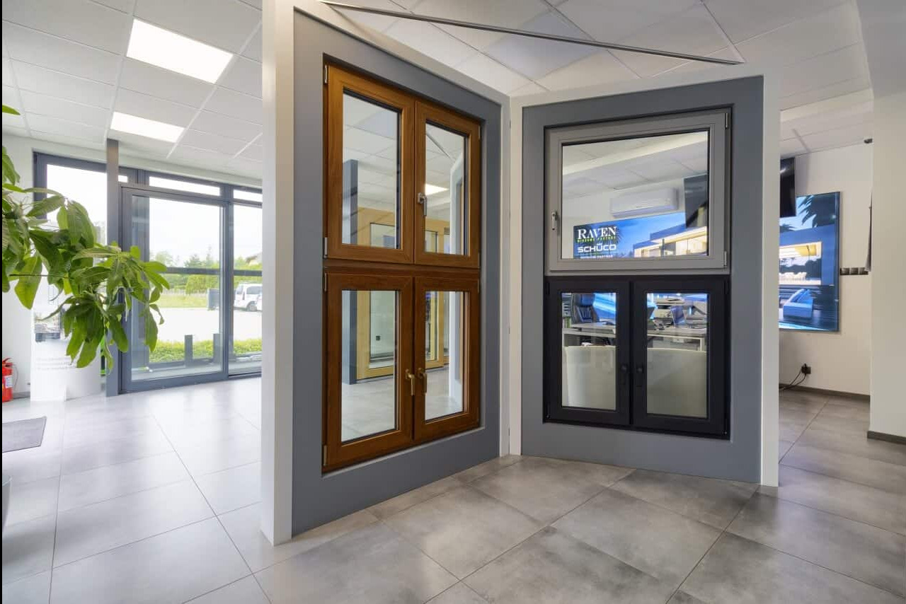 Ekspozycja okien PCV: brązowe drewniane okno krosnowe i czarne okno z szybą zespoloną na szarym tle w salonie wystawowym z rośliną i oknami przesuwnymi.