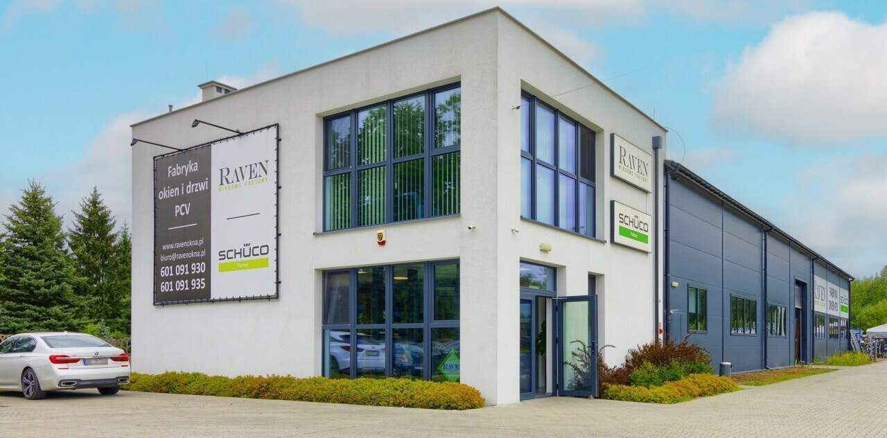 Budynek firmy Raven, producenta okien i drzwi PCV. Widoczne okna z szprosami i drzwi. Na elewacji logo firmy i marki Schüco. Zaparkowany samochód BMW.